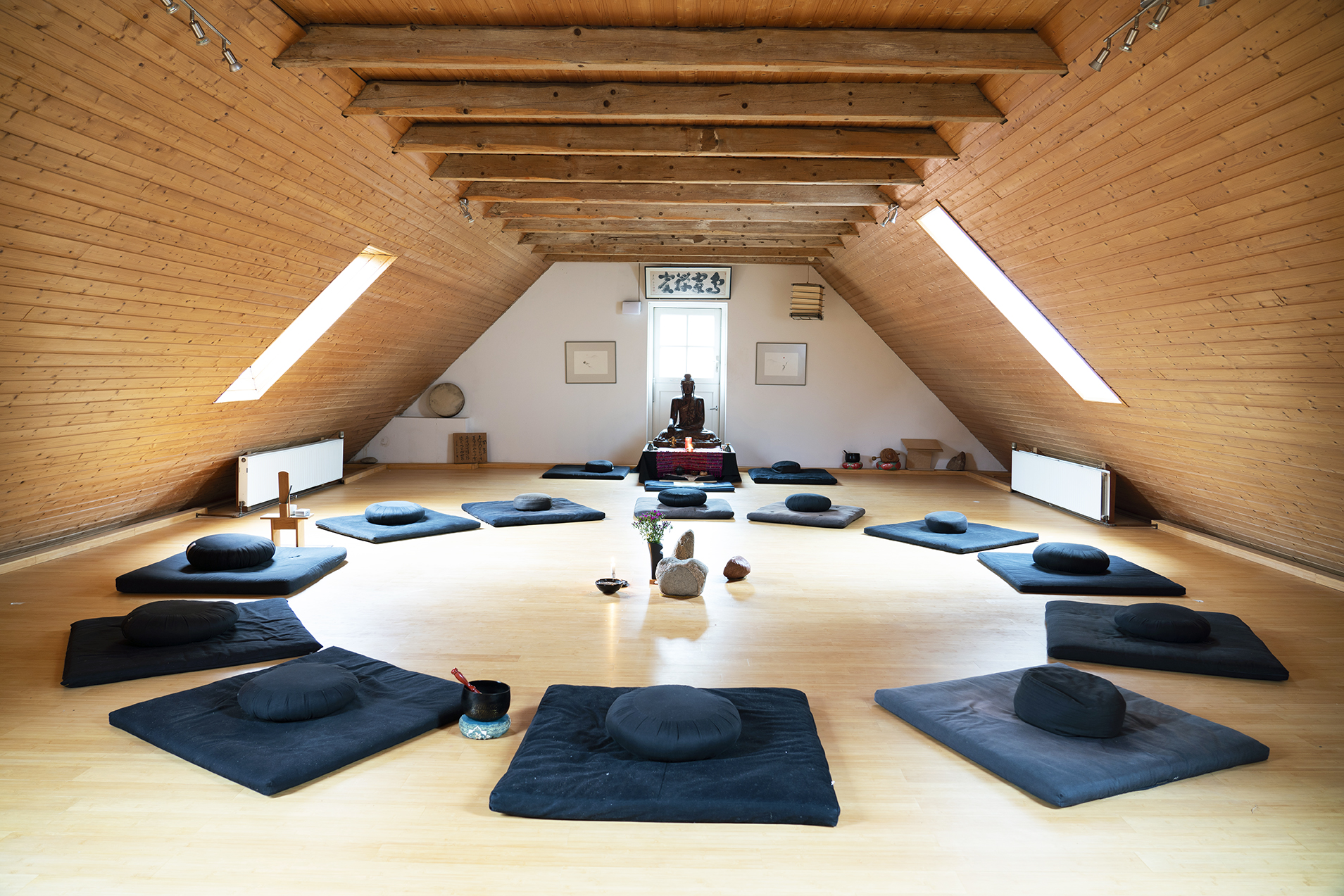 Im Zendo-Raum im Lebensgarten Steyerberg liegen Meditationskissen in einem Kreis angeordnet. Am Kopfende des Raumes steht eine Buddha-Figur.