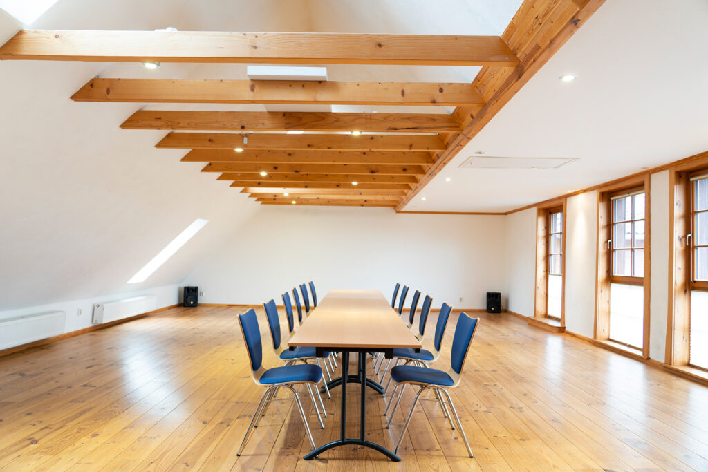 In einem schönen hellen Seminarraum mit Holzboden und Dachbalken steht ein langer Tisch mit zwölf Stühlen.