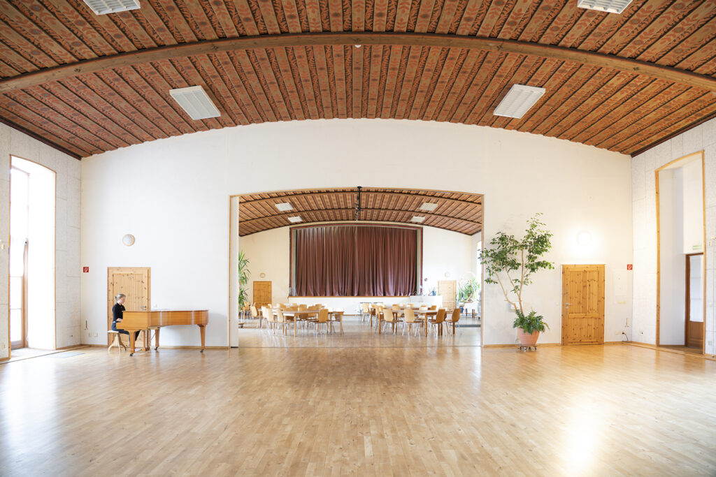 Sehr großer heller Raum mit Holzboden und gewölbter Decke. Blick in den Speisesaal und die Bühne in den Räumlichkeiten der CAIA academy.