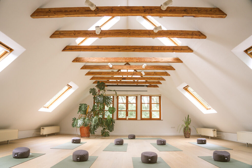 Schöner heller Yogaraum mit Dachschrägen und Holzbalken mit Blick in den Wald des Lebensgartens Steyerberg